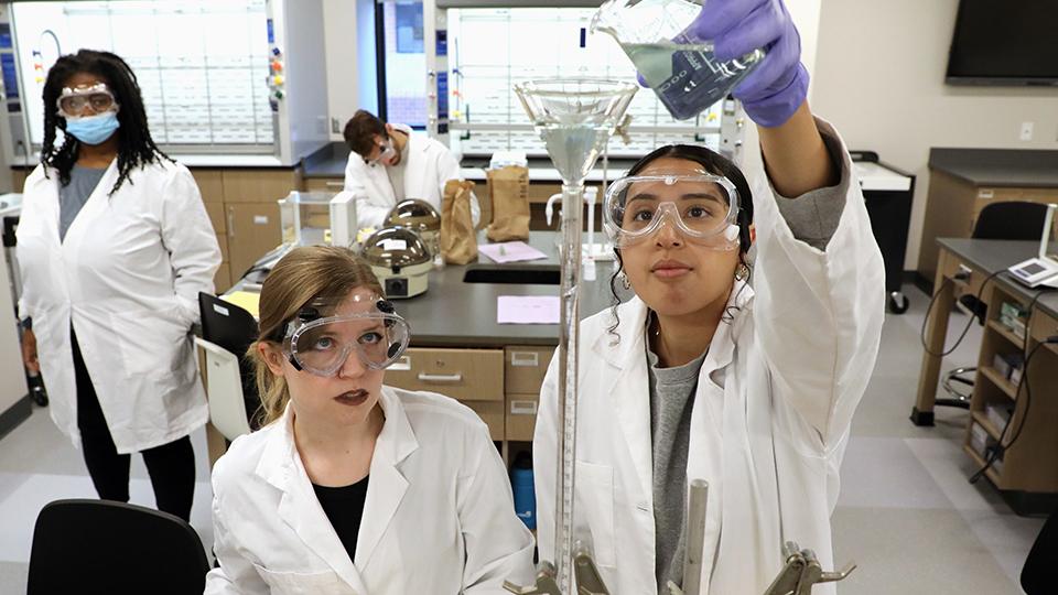 two jccc students in lab coats and safety gear pour a beaker of liquid into a tall funnel while other students work in the lab in the background