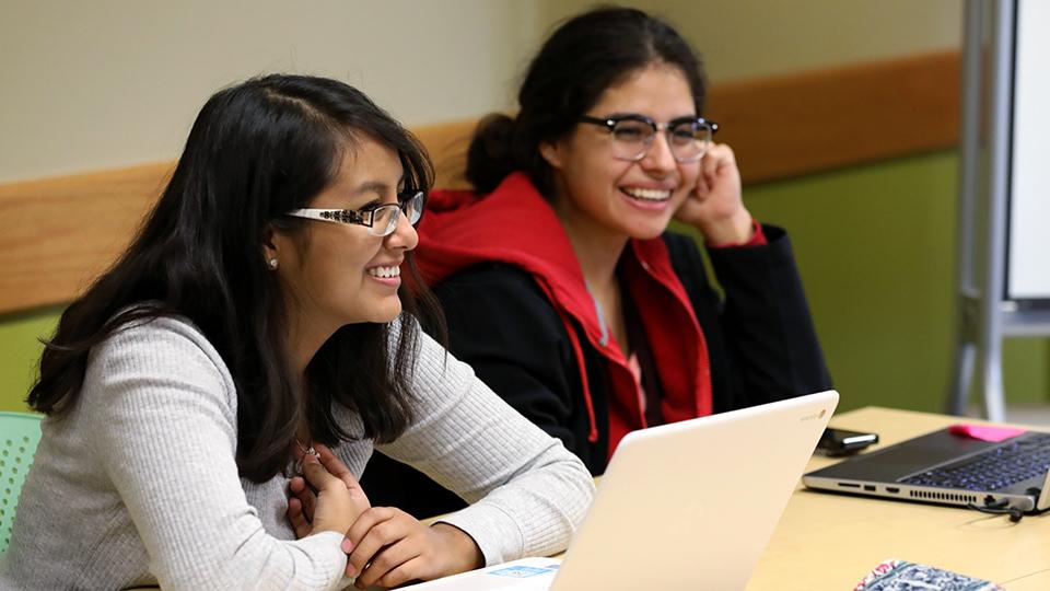 Two students at a table with laptops.