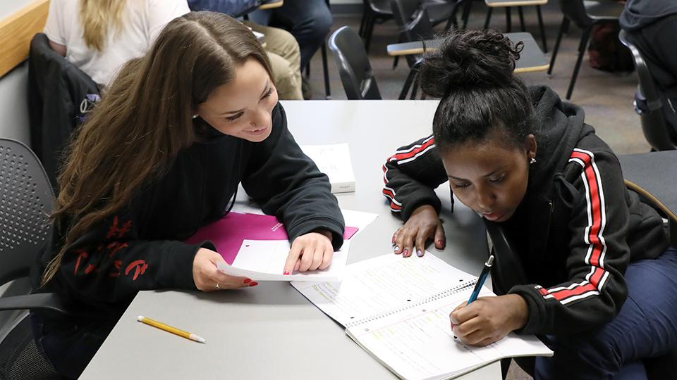 One student writes in a notebook while another looks over the work.