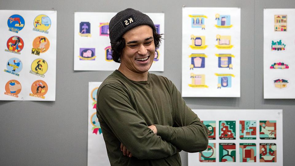 Smiling student standing in front of a display board of student graphic design work