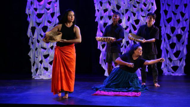 performers on stage - a woman carrying a basket walks past three other people, two of whom are standing holding baskets and another who is on her knees with arms outstretched