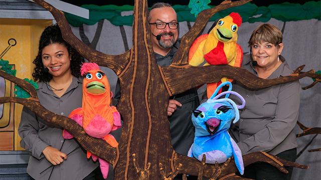 3 smiling puppeteers wearing microphones hold their colorful puppets up on a tree trunk